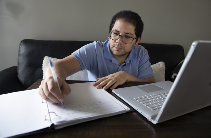 Díaz sits on a couch leaning over a table in front of him while taking notes on a sheet of paper in an open binder next to his laptop, both of which are sitting on the table.