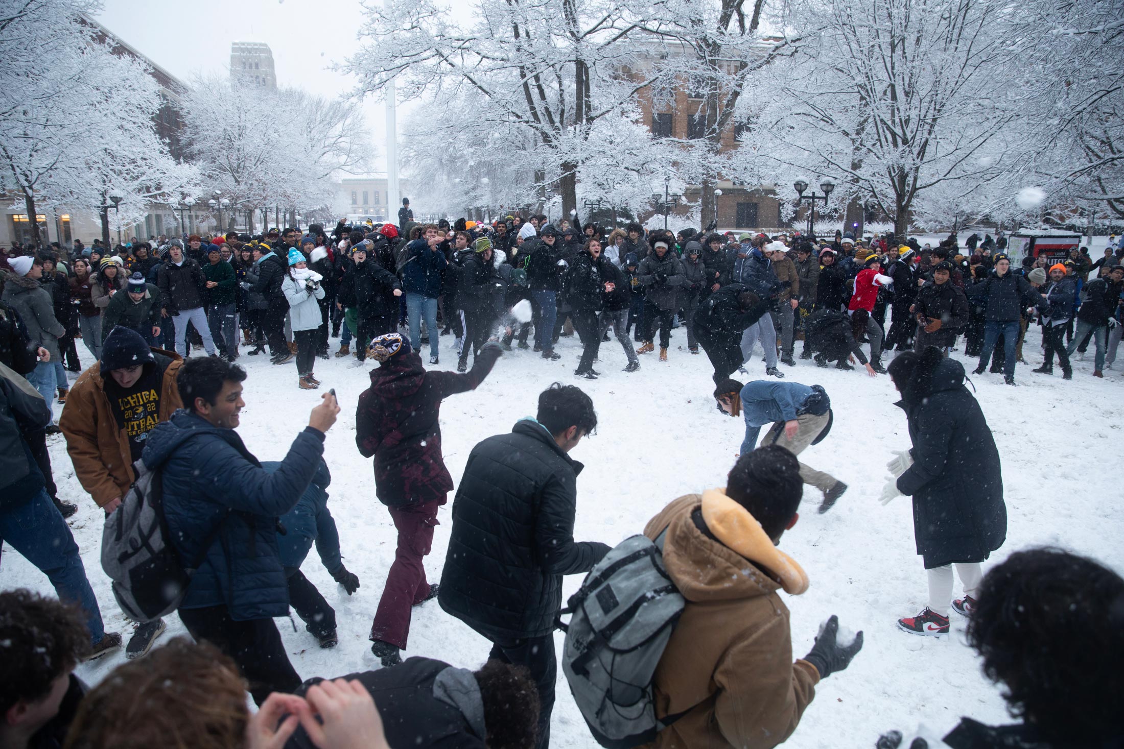 A large group of people participate in an outdoor snowball fight surrounded by snow-covered trees and buildings. Snowballs are being thrown and are in mid-air.