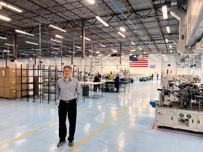 Albert Shih stands in a clean, mostly empty factory floor. It's brightly lit and machinery and workers in gear can be seen in the background