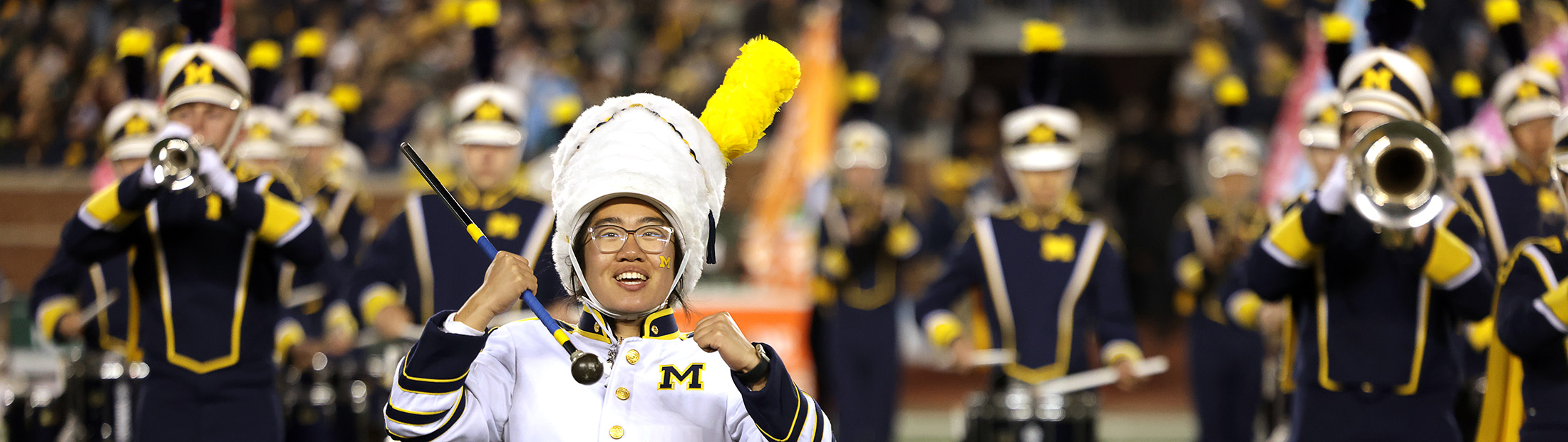 Rachel Zhang in her drum major uniform with fists partially raised while surrounded by other marching band members playing their instruments on the field at Michigan Stadium under the lights.