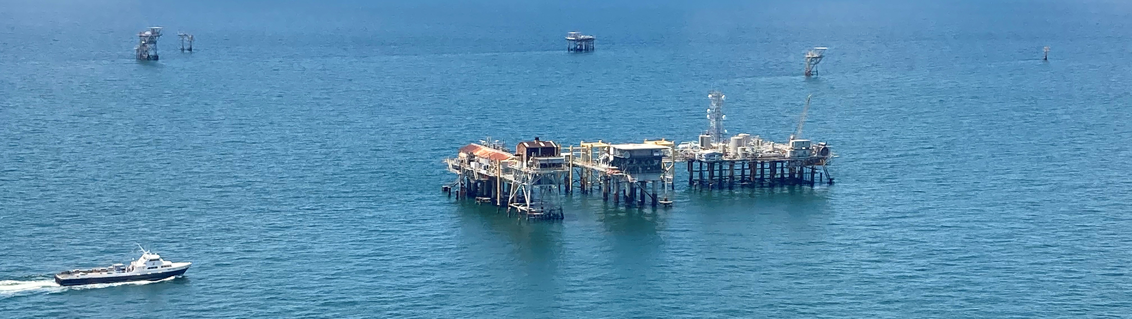 An offshore drilling complex sits over blue water. A large boat is emerging from the left.