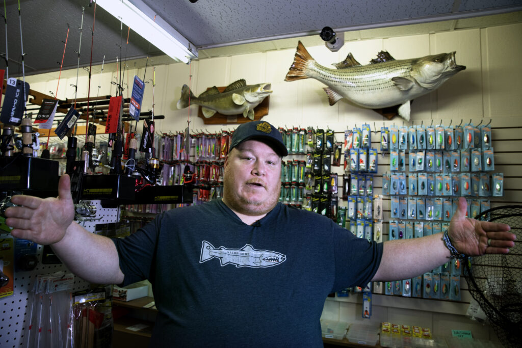 Roger Byrne, who works at Wellman’s Bait & Tackle, spreads his arms apart as if to show a large distance. Behind him is a wall of fishing gear and equipment, as well as some mounted fish trophies.