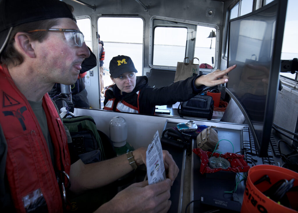 Standing in the cabin of their research vessel, Katie Skinner points to a monitor while a discussing wreck findings with a colleague 