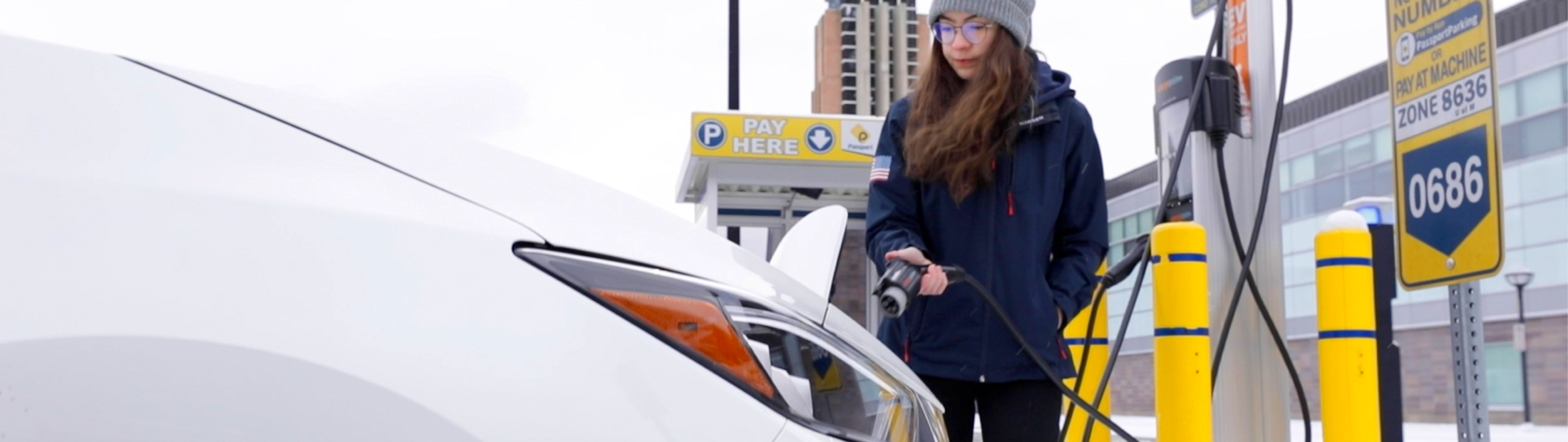 A woman preparing to plug in an electric vehicle. She's wearing a winter coat and hat. In the background, you can see Lurie Tower.