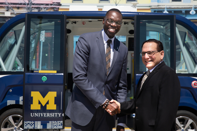 Two men smiling and shaking hands. Behind them is a U-M autonomous shuttle