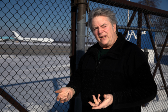 A man in a black winter jacket stands beside a rusty fence making a puzzled or perhaps bewildered face. In the distance you can see a large airplane. The ground has a small amount of snow on it.