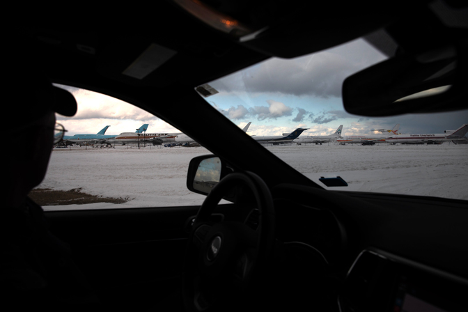 A man sits in the drivers seat of his car and looks at retired planes behind a rusty fence. The Inside of the car is almost too dark to make out details and the partly cloudy sky add extra dreariness to the image.