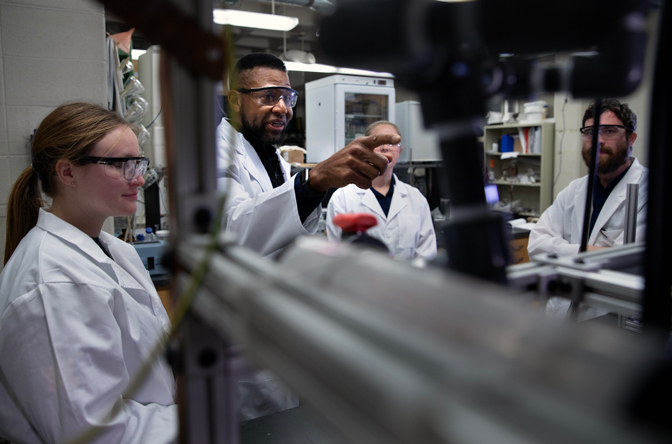 John Foster and three graduate student researchers in white lab coats and safety glasses look at lab equipment during a demonstration. The subjects of the photo are partially obfuscated by lab equipment.