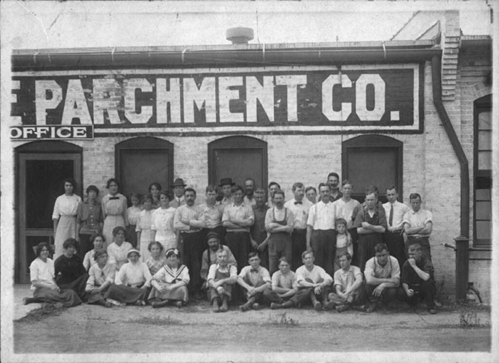 A black and white photo showing over two dozen men and women from the paper mill standing and sitting outside the building. Above them you can partially make out the words "Parchment Co." 
