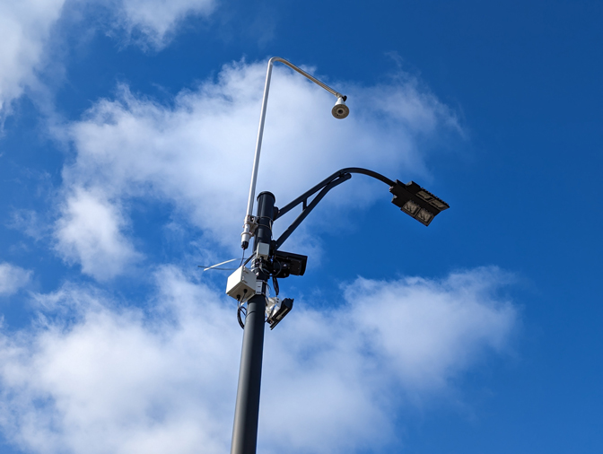 a series of wires and bits of equipment are mouonted to a light pole. Beyond the pole you can see clouds and a blue sky.