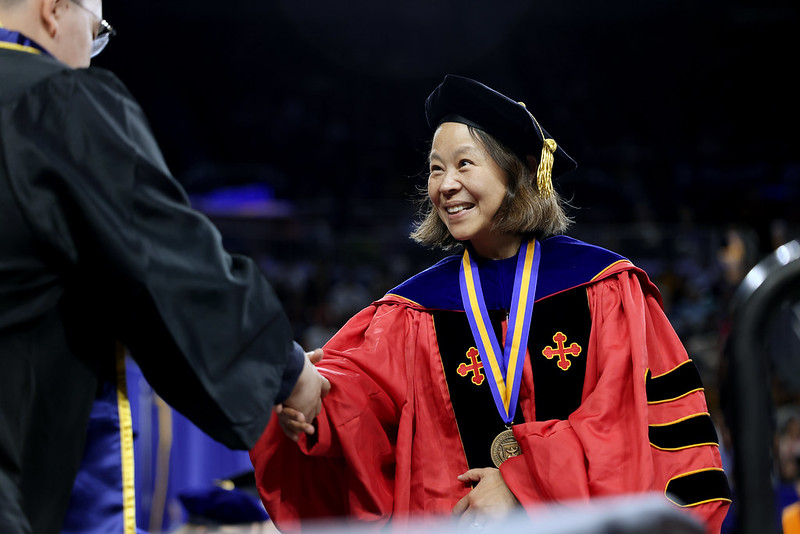 Mingyan Liu is wearing a red cap and gown for faculty. She is smiling and shaking hands with a student (back to the camera) who is wearing the traditional black graduation cap and gown 