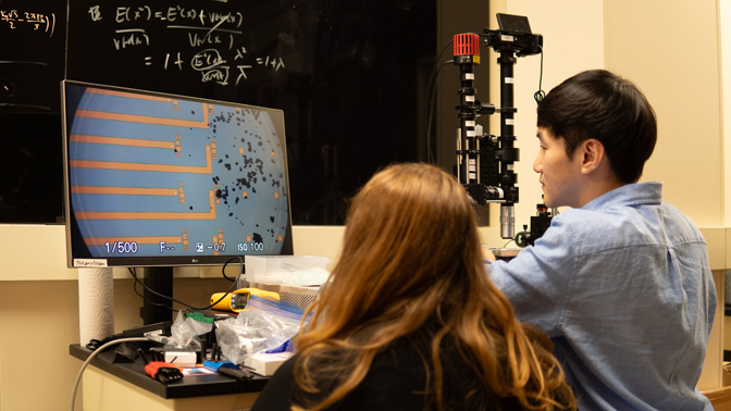 A monitor shows a circle with orange lines, which are microelectrodes, stretching towards the battery particles, which look like black dots on the screen. Two students observe the screen
