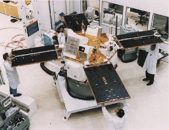 The ACE space probe appears as a cylinder that is slightly taller than the four scientists standing around it. Each scientist is wearing a white lab coat and is holding a solar panel extending from the space probe. The solar panels look like flat rectangles with a black surface and gold edge. The space probe is capped in reflective, gold foil and various boxy, white instruments.