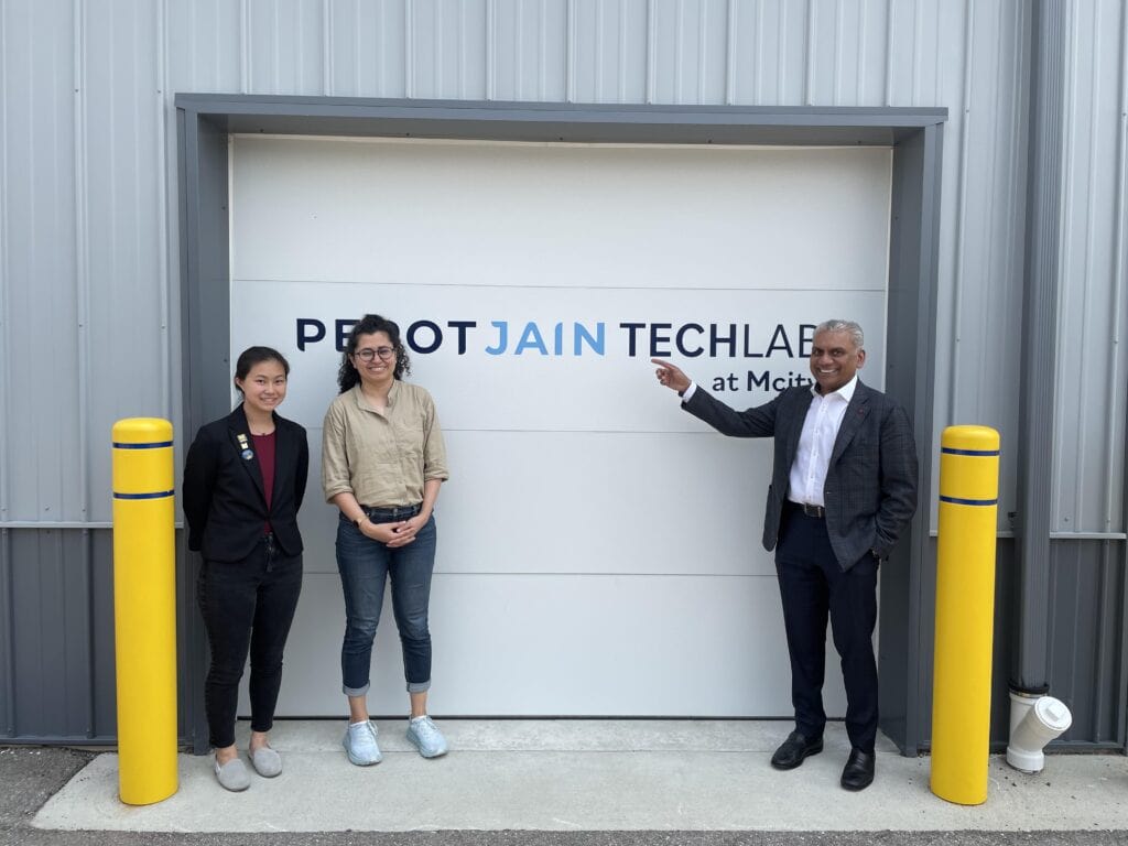 Two women and a man stand in front a building sided in gray corrugated metal, with the man pointing at the "PEROTJAIN TECHLAB at Mcity" logo on the white sectional garage door.