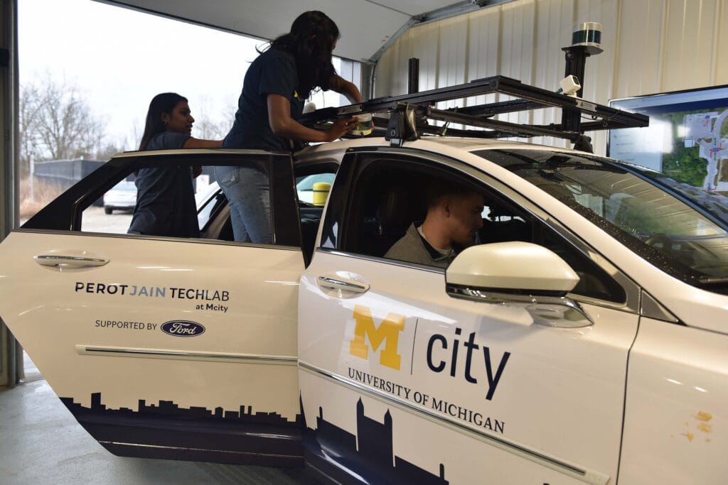 One woman stands just inside an open car door, manipulating something on a frame mounted to the roof of a white car labeled with Perot Jain TechLab, Ford, Mcity and the University of Michigan. Another woman stands behind her, appearing to help, while a man sits in the passenger seat, looking down and toward the driver's side.