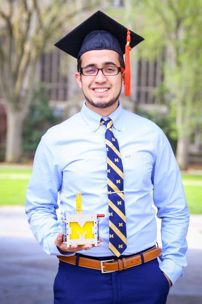 A man stands in a blue button shirt, navy pants and blue and yellow striped tie. He's wearing a graduation cap and holding a Block M.