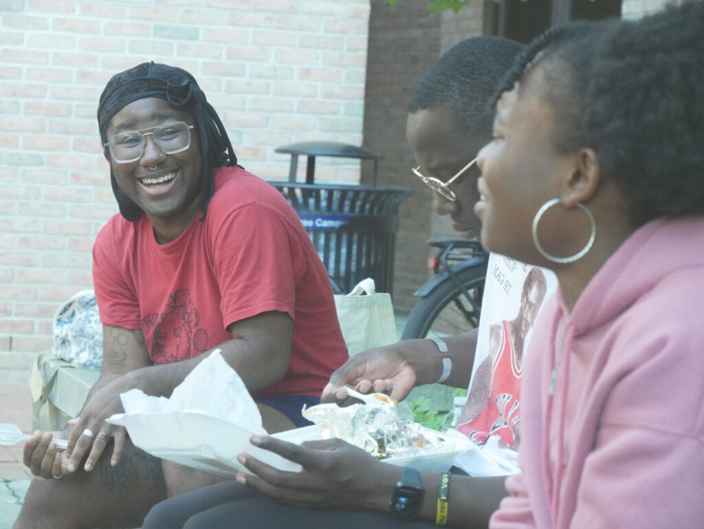 Three students are sitting on a brick wall. On the left is a smiling, bespectacled student wearing a red shirt. Another student wearing glasses sits in the center, and they are eating food on a styrofoam tray in their lap. The rightmost student is wearing a pink hooded sweater and is leaning around the center student to chat with the leftmost student.