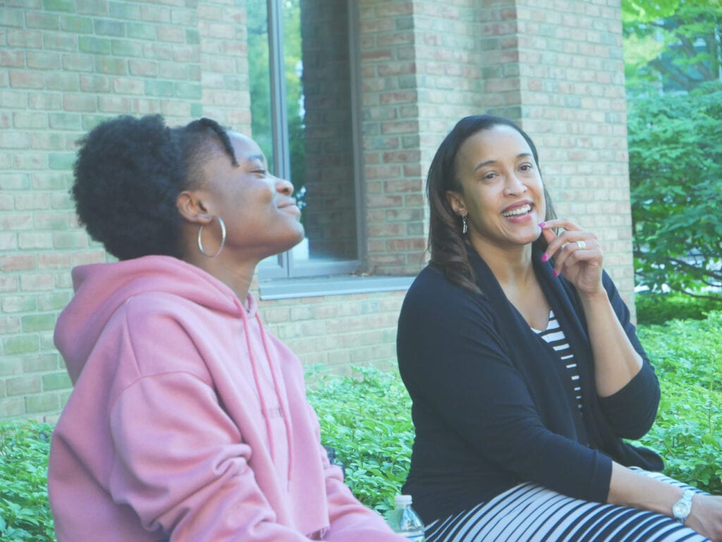 A woman wearing hoop earrings and a pink, hooded sweater sits outside on a bench with another woman wearing a black cardigan over a dress with black and white stripes. Mondisa, on the right, is stroking her chin and smiling, while Ngozi, on the left, appears to be nodding in agreement.