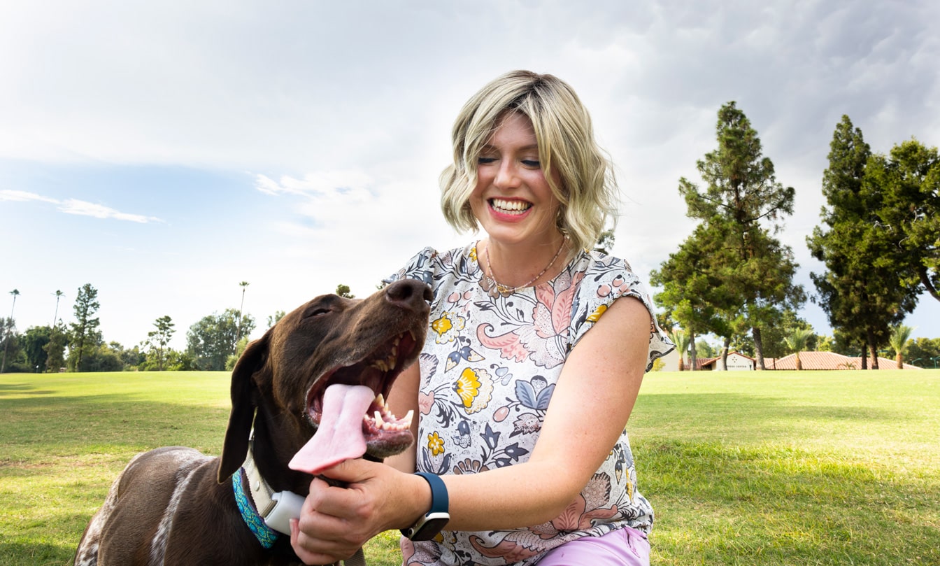 A woman smiles while petting her chocolate lab in a park on a sunny day