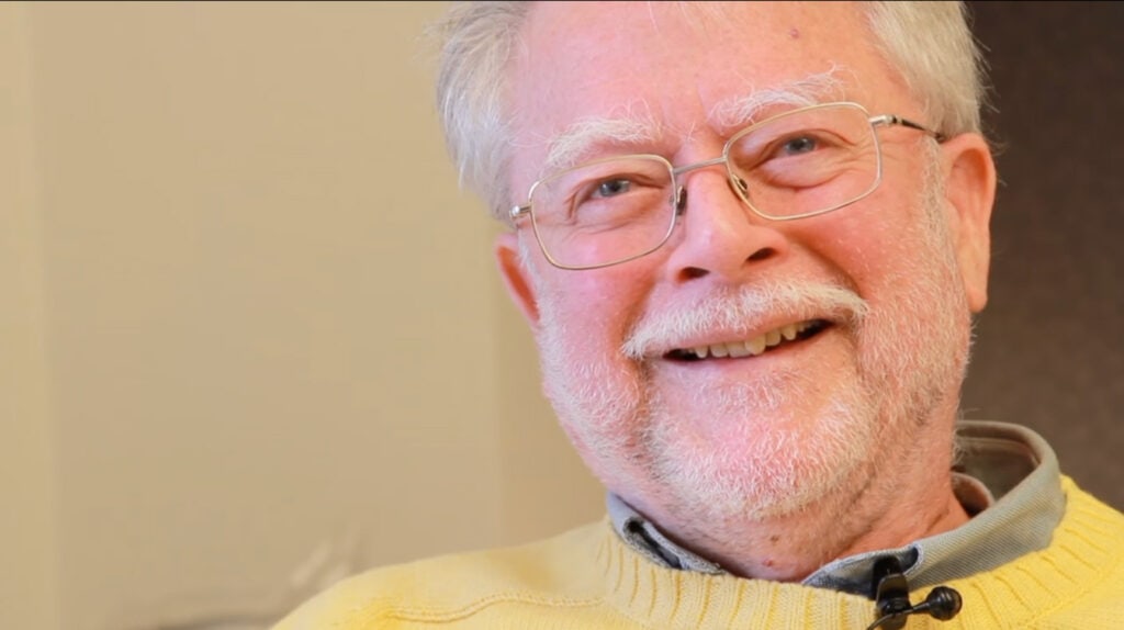 A man with a white hair and a beard smiles in a yellow sweater