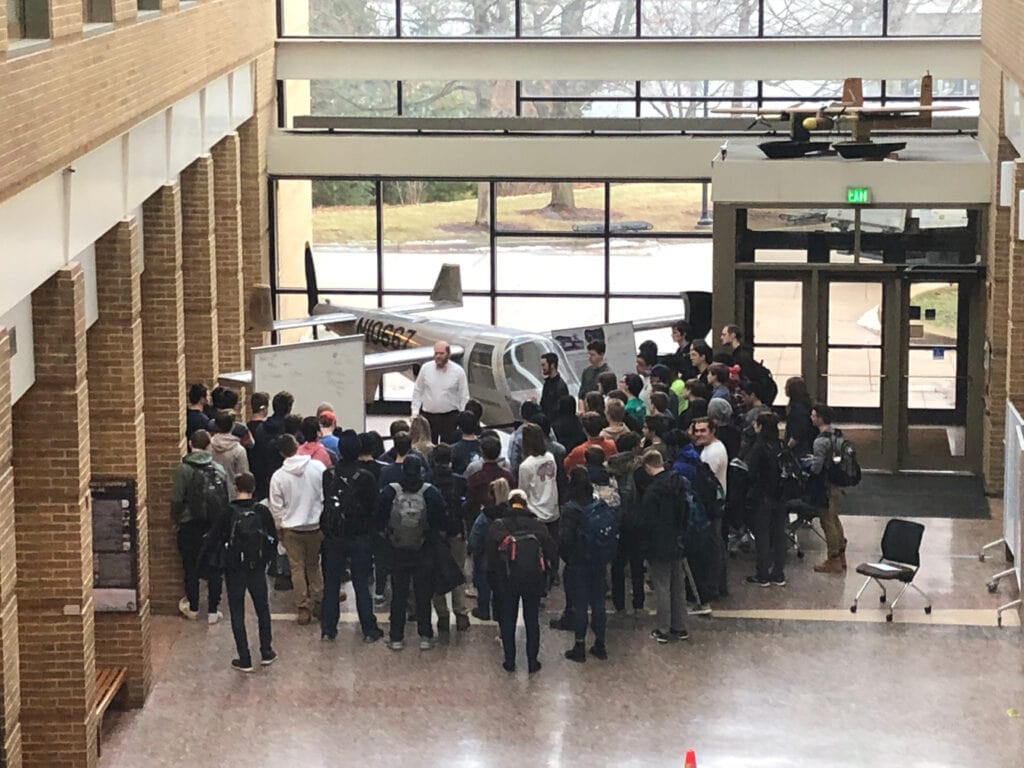 A group of around 50 students gathered in front of a small white aircraft and a whiteboard. A man in a white shirt stands in front of the whiteboard and aircraft and faces the group of students.