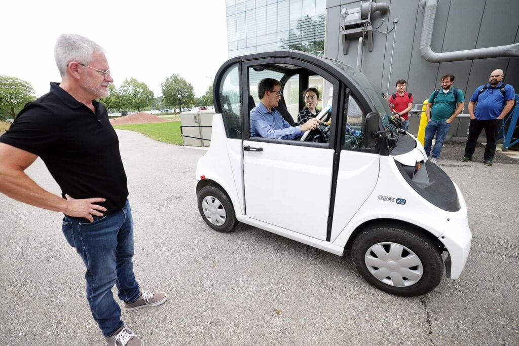 A mans with his hands on his hips watches as two people sit inside a small vehicle