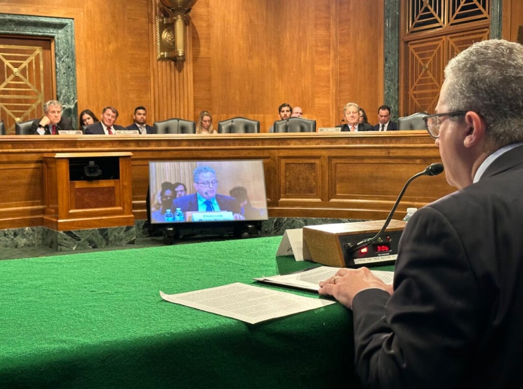 A man in a suit, Professor Wellman, sits at a table covered with green felt. He is speaking into a microphone with his back facing the frame. In front of Wellman is a TV monitor that is displaying his face. Behind the monitor, the Senate committee members are sitting at a raised bench.