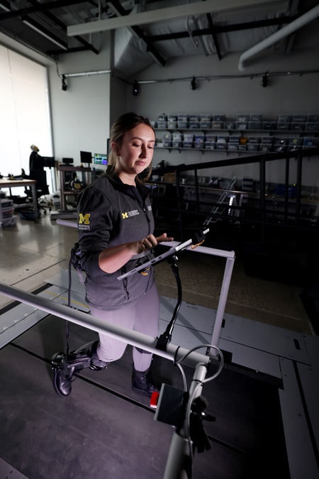 A woman walks on a treadmill while wearing the exo-leg. She adjusts controls on an iPad with one hand.