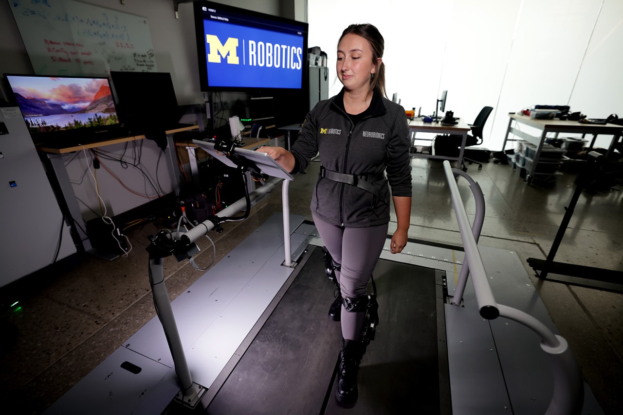 A woman walks on a treadmill while wearing the exo-leg. She adjusts controls on an iPad with one hand.