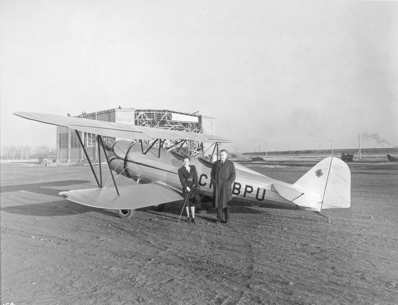 Black and white photo pf Elsie MacGill, holding a cane, on the left and a man on the right stand in front of a two seater aircraft on a dirt runway.