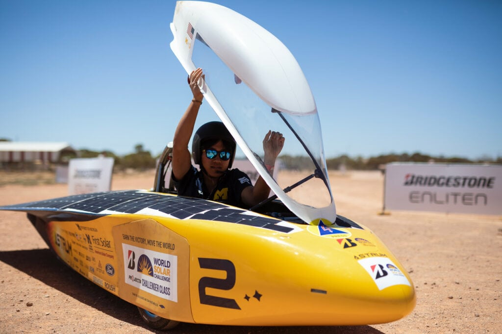 A driver is lifting Astrum's round, white hood so that he can lower himself into the driver's seat. He is wearing a helmet with an attached microphone and sunglasses. Astrum's body is yellow and bullet shaped and is covered in logos from the team's various sponsors.