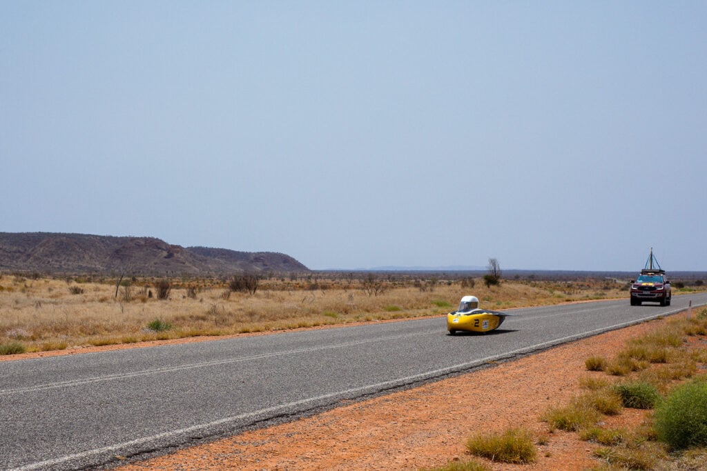 Astrum advances along a road in the Australian outback. The sand surrounding the gray road is a red-brown color. Astrum is shaped like a yellow bullet. A round, white hood covers the driver's seat. A maroon chase car with an antenna on its hood follows Astrum. 