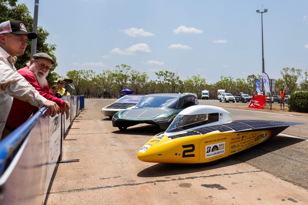 Astrum is arranged in front of two other solar cars. Astrum's body is yellow and bullet shaped while the cockpit is white. The second car in line is a green catamaran, with solar panels on the hood. It sits in front of the third car in line is white with solar panels on the hood. Onlookers observe from behind a fence to the left of the cars.