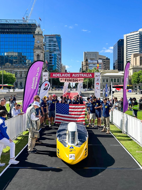 The Michigan solar car team stands behind their yellow car, Astrum, at the ceremonial finish line in Adelaide, Australia. Tall buildings are visible in the background, and a red banner reads "Adelaide Australia" over the. The entire team is wearing maize and blue and are holding up the American flag.