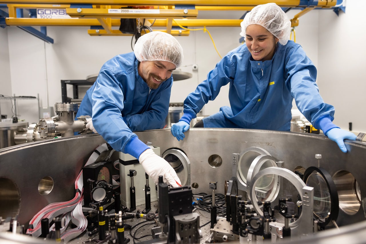 Two researchers in full lab gear lean over a cylindrical chamber. One researcher leans in to make an adjustment.