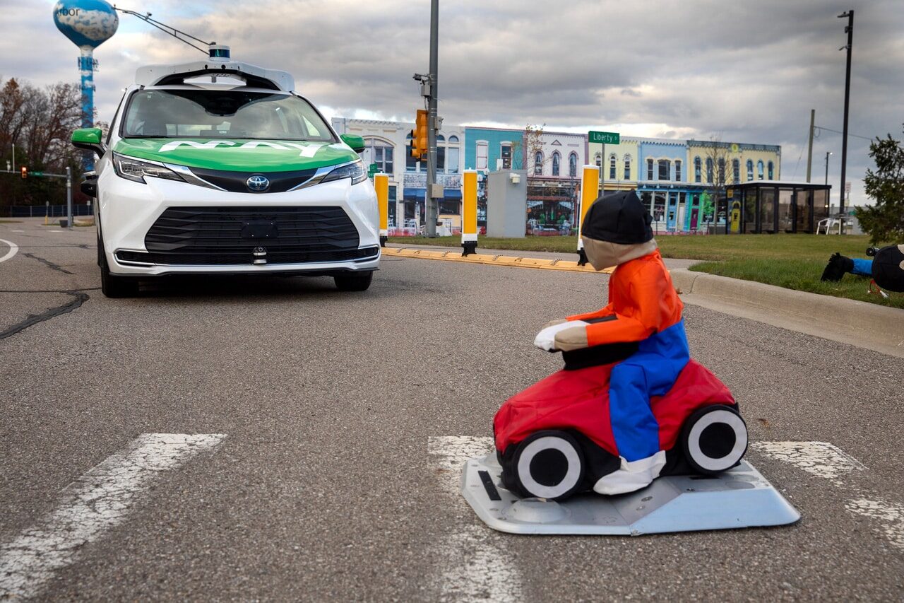 A test dummy child in a cross walk. Behind it, a green and white autonomous van stands still.