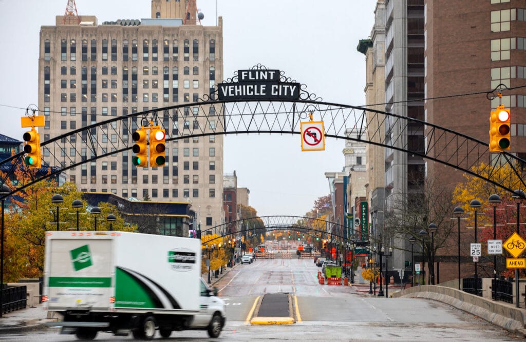 An enterprise truck turns down a double lane road in downtown Flint. Over the roadway, a sign reads 'Flint: Vehicle City'