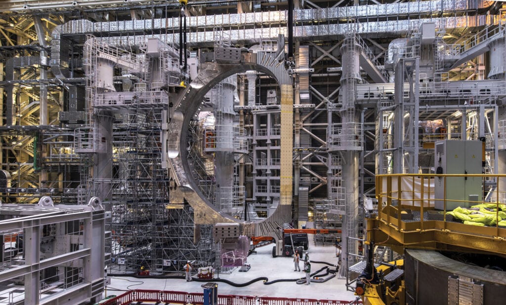 Several stories high Tokamak under construction. Around it you can see scaffolding to make assembly easier. At the base of the construction, you can see workers. By scale, Tokamak appears to be dozens of feet tall.