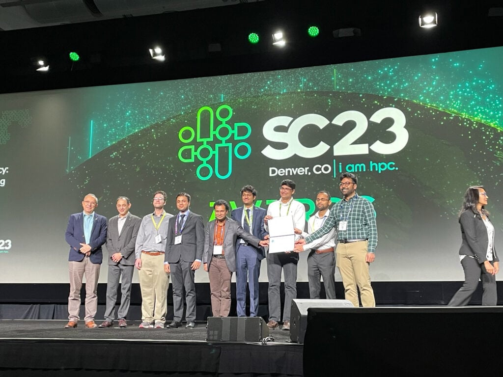 Nine men stand onstage with Vikram Gavini holding a  a stage-size screen reading SC23 | Denver, CO | i am hpc behind them.