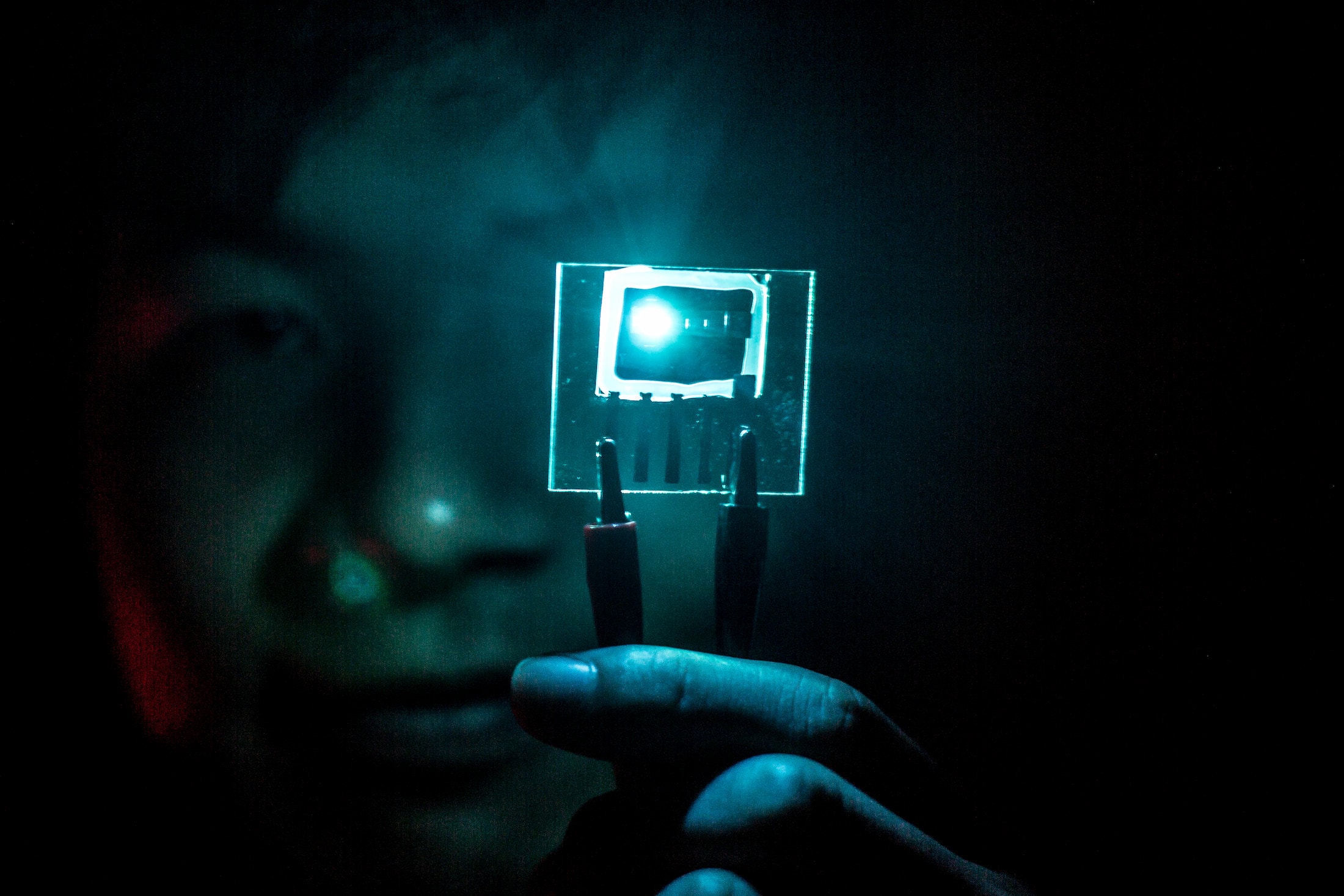 A researcher uses forceps to hold up a small chip in a dark room. The chip contains a blue PHOLED, which lights up the surrounding area. The lighting makes the researcher's hands and face appear a blue-green color.