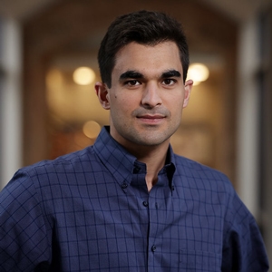 Mojtaba Akhavan-Tafti smiles in a portrait photo. He's wearing a plaid blue shirt and is standing in a large hallway