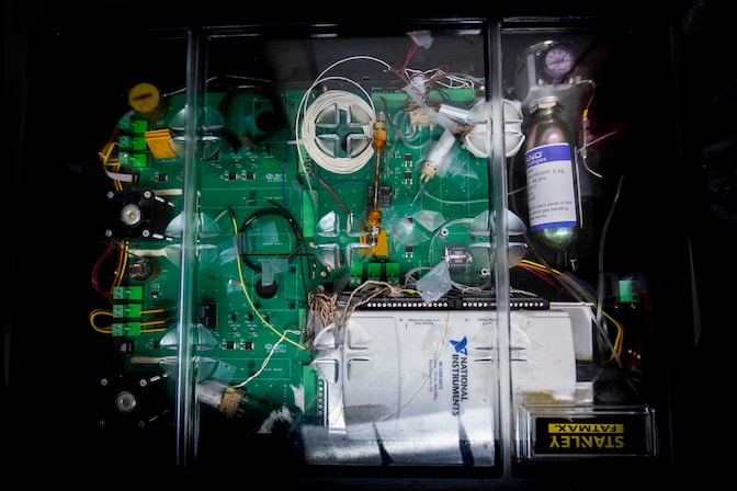 A green board is criss-crossed by wires and tubes, and a white box occupies the lower-right corner. The set-up includes a pressurized bottle with a dial gauge.