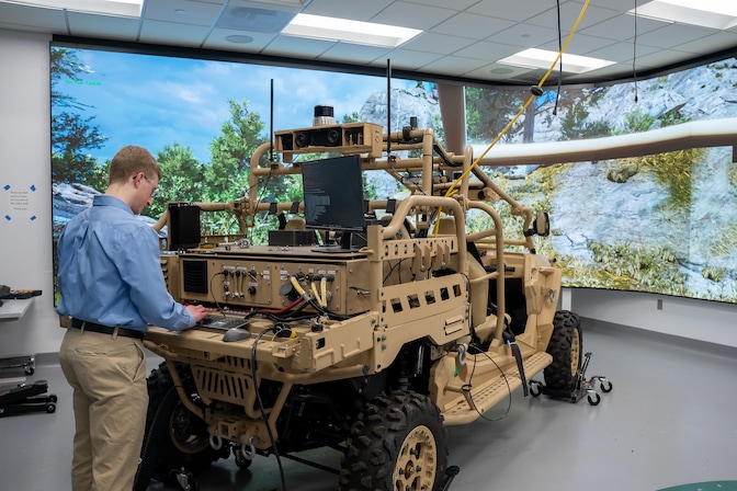 A researcher works on a desktop attached to the back of one of the military off-road vehicle model simulators. A floor to ceiling projector screen of a outdoor nature scene with pine trees is wrapped around the walls in front of the vehicle to enhance the simulation experience.