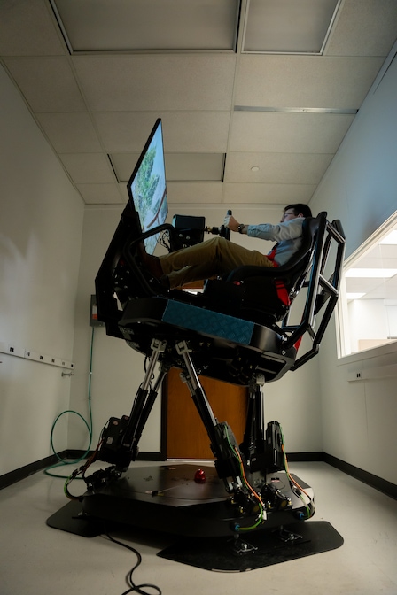 PhD student James Baxter sits in an ARC simulator that sits several feet off the ground held by four metal rods, and consists of of a large screen displaying outdoor scenery, hand controls, and a passenger seat.