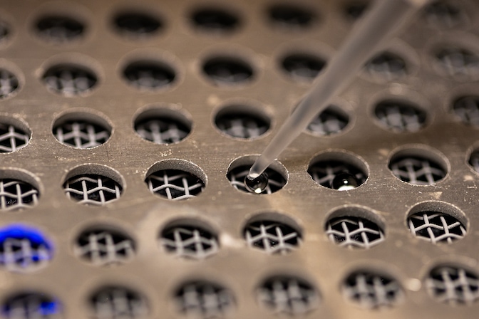 A neat grid of holes are drilled into a metal plate, which is covering a gray screen mesh. The screen is visible beneath the holes of the plate. A plastic pipette tip deposits small beads of water into several wells.