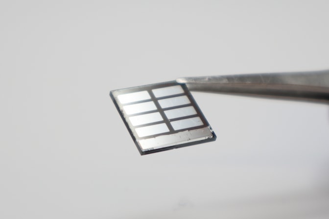 A pair of forceps hold a perovskite solar cell, which looks like a thin, square panel. The cell's electrodes look like silver rectangles on the cell's surface, and the surrounding black surface is the perovskite film.