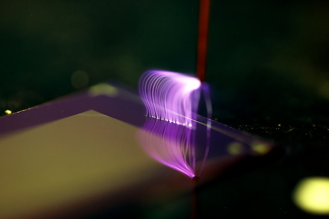 Bright purple, lightning-like plasma arcs from the tip of the metal wand onto the blue chip. The plasma stands in stark contrast with the dark background and illuminates the wand, causing them to appear redder than usual.