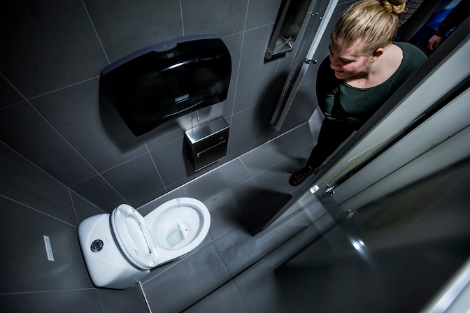 A student examines a source-separating toilet in a stall. The toilet has a split bowl and is designed to send solid waste to a wastewater treatment plant, but route urine to a holding tank.