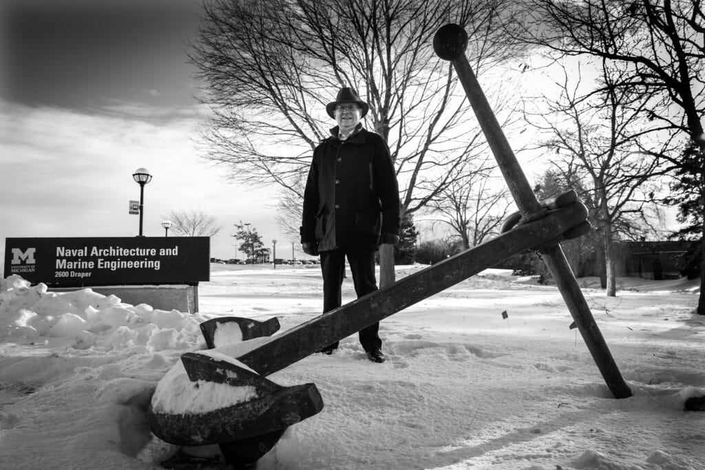 On a snowy winter day, Donald Winter stands next to the sign for the Naval Architecture and Engineering building on U-M’s North Campus. In front of him is a large ship’s anchor that’s on display, its iron crosspiece taller than he is.