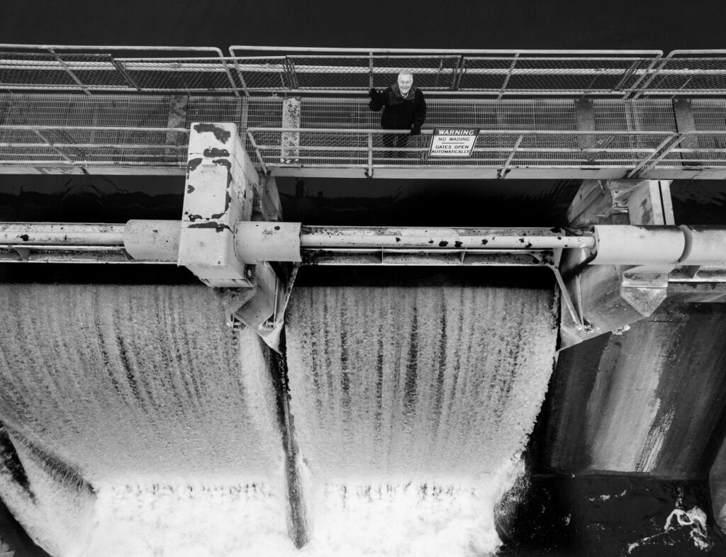 Standing on a walkway that spans a small hydroelectric dam, Glen Daigger smiles up toward an aerial camera as water cascades down the dam’s spillway.